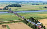 245 024 schiebt den RE 11017 nach Hamburg-Altona über die Eiderbrücke bei Friedrichstadt, die gerade renoviert wird. Aber wo ist denn nun 012 104? Das geübte Auge findet den Sonderzug mit der Dampflok in der Kurve hinter dem Wäldchen. Dort wartet der Zug die Zugkreuzung des RE ab. (20.07.2024) <i>Foto: Joachim Schmidt</i>