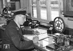 Der Fahrdienstleiter im Bahnhof Königsmoor an der Strecke Bremen - Hamburg mit dem damals zur Nachrichtenübermittlung üblichen Morse-Fernschreiber, umgangssprachlich auch "Telegraph" genannt. Er schreibt hier die soeben empfangene Zugmeldung in das Zugmeldebuch. Die Fahrdienstleiter mußten damals noch das "Morsen" beherrschen, um mit Hilfe der Morsetaste (rechts unten im Bild) die Zugmeldung senden zu können, bzw. die empfangene Zugmeldung vom Papierstreifen lesen zu können. Das Wort "ZUG" sieht in Morseschrift z.B. so aus:   _ _..   .._    _ _. Links im Bild sieht man einen zweiten Fernschreiber, den sogenannten Bezirkstelegraphen. Dieser ist mit allen Stationen der Strecke gleichzeitig verbunden. (30.07.1954) <i>Foto: Walter Hollnagel</i>