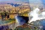 Ein Zug auf der 1905 eröffneten Victoria Falls Bridge, 128 m über dem Sambesi, der hier die Grenze zwischen Rhodesien (heute Simbabwe) und Zambia bildet. Der Sambesi fließt von rechts nach links und führte zum Zeitpunkt der Aufnahme relativ viel Wasser, sodass die Gischtwolken, die der Wasserfall produziert deutlich sichtbar sind. Die Wassermassen des Sambesi ergießen sich hier auf einer Breite von 1708 m in eine quer zum Flusslauf liegende, 110 m tiefe und kaum mehr als 50 m weite Schlucht mit steilen Felswänden aus Basalt. Damit sind die Victoriafälle der breiteste durchgehende Wasserfall der Erde. Zum Ende der Regenzeit im Februar und März schießen bis zu 10.000 m³/s Wasser in die Tiefe. Zum Ende der Trockenzeit, in den Monaten September und Oktober, kann die Wassermenge jedoch auf nur 170 m³/s schrumpfen. Dann bleiben von der sonst tosenden Flut nur einige wenige Rinnsale übrig.  (26.07.1975) <i>Foto: Herbert Vaupel</i>