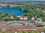 Vor dem Bw Neustrelitz und der Kulisse des Glambecker Sees schiebt eine 143 den RE 13056 nach Stralsund aus dem Bahnhof Neustrelitz. (14.06.2025) <i>Foto: Joachim Schmidt</i>
