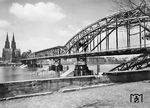 Nachdem am 6. März 1945 die Brückenpfeiler der Hohenzollernbrücke in Köln durch Pioniere der sich zurückziehenden Deutschen Wehrmacht gesprengt wurden, konnte ab 1948 wieder ein zweigleisiger Betrieb mit einem provisorischen SKR-Stahlüberbau über die Brücke aufgenommen werden. Als Ersatz für die zerstörte Straßenbrücke auf der Südseite wurde lediglich ein Geh- und Radweg angesetzt. Auf der Deutzer Seite lag noch 1948 ein Rest eines alten Brückenüberbaus im Rhein. (1948) <i>Foto: Fischer</i>