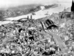 Die Stadt Köln wurde im Laufe des 2. Weltkriegs 262 mal bombardiert, mehr als jede andere deutsche Stadt. Mit der "Operation Millennium" war Köln in der Nacht vom 30. auf den 31. Mai 1942, Ziel der Royal Air Force (RAF), die hier erstmals über 1000 Bomber gleichzeitig einsetzte. Rund 20.000 Kölner starben durch die Luftangriffe. Der verheerendste Angriff erfolgte in der Nacht auf den 29. Juni 1943, bei dem etwa 4300 Menschen starben und rund 230.000 obdachlos wurden. Am 2. März 1945 griff die RAF Köln zum letzten Mal mit 858 Bombern in zwei Phasen an. Im Rahmen der "Operation Lumberjack" wurde wenige Tage später der linksrheinische Teil Kölns von der 1. US-Armee eingenommen, am Abend des 6. März 1945 war der Krieg in Köln beendet. Der deutsche Rundfunk meldete hierzu: "Der Trümmerhaufen Köln wurde dem Feinde überlassen." Im Frühjahr 1945 lebten nur noch 40.000 Menschen in Köln; vor dem Krieg waren es 768.000. Links des Hauptbahnhofs liegt die (von den Deutschen) gesprengte Hohenzollernbrücke im Rhein, rechts daneben die Deutzer Hängebrücke, ganz oben die Kölner Südbrücke.  (24.04.1945) <i>Foto: Pressefoto ACME</i>