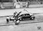 Erwischt: Während der Fahrer des VW-Käfers versucht, sein Malheur der umgefahrenen Mülltonnen an der Stoßstange seines fahrbaren Untersatzes zu beseitigen, darf die Ehefrau die Folgen zusammenkehren und hofft, dabei nicht beobachtet zu werden. (1951) <i>Foto: Slg. Wolfgang Löckel</i>